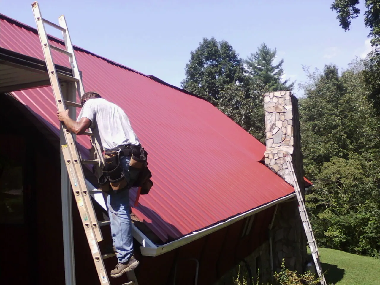 Roofer on a ladder beside a red metal roof and stone chimney — Soffit & Fascia Repair by Roofing Brothers in Panama City Beach